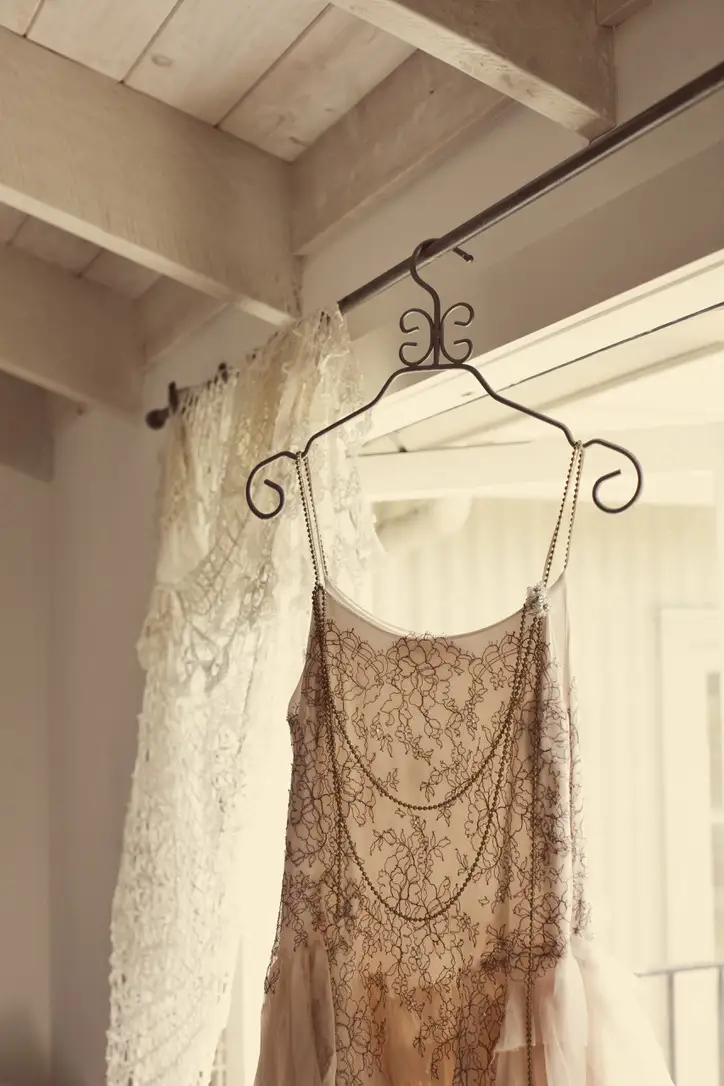 A delicate lace dress with beaded chains hangs on an ornate metal hanger in a softly lit room with exposed wooden beams and a lace curtain by a window.