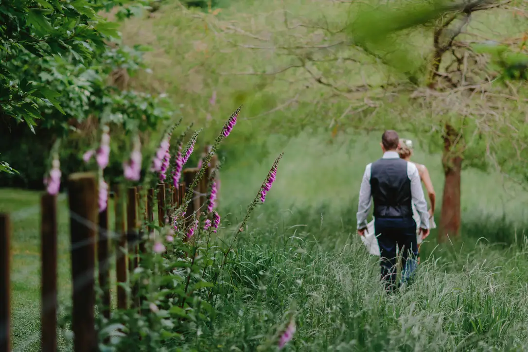 A couple walks away down a grassy, flower-lined path surrounded by lush greenery, with tall pink wildflowers in the foreground and trees arching overhead.