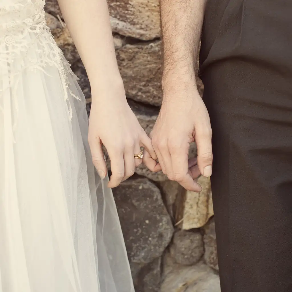A bride in a white lace dress and a groom in a dark suit gently hold hands, showing their wedding rings. The background features a stone wall.