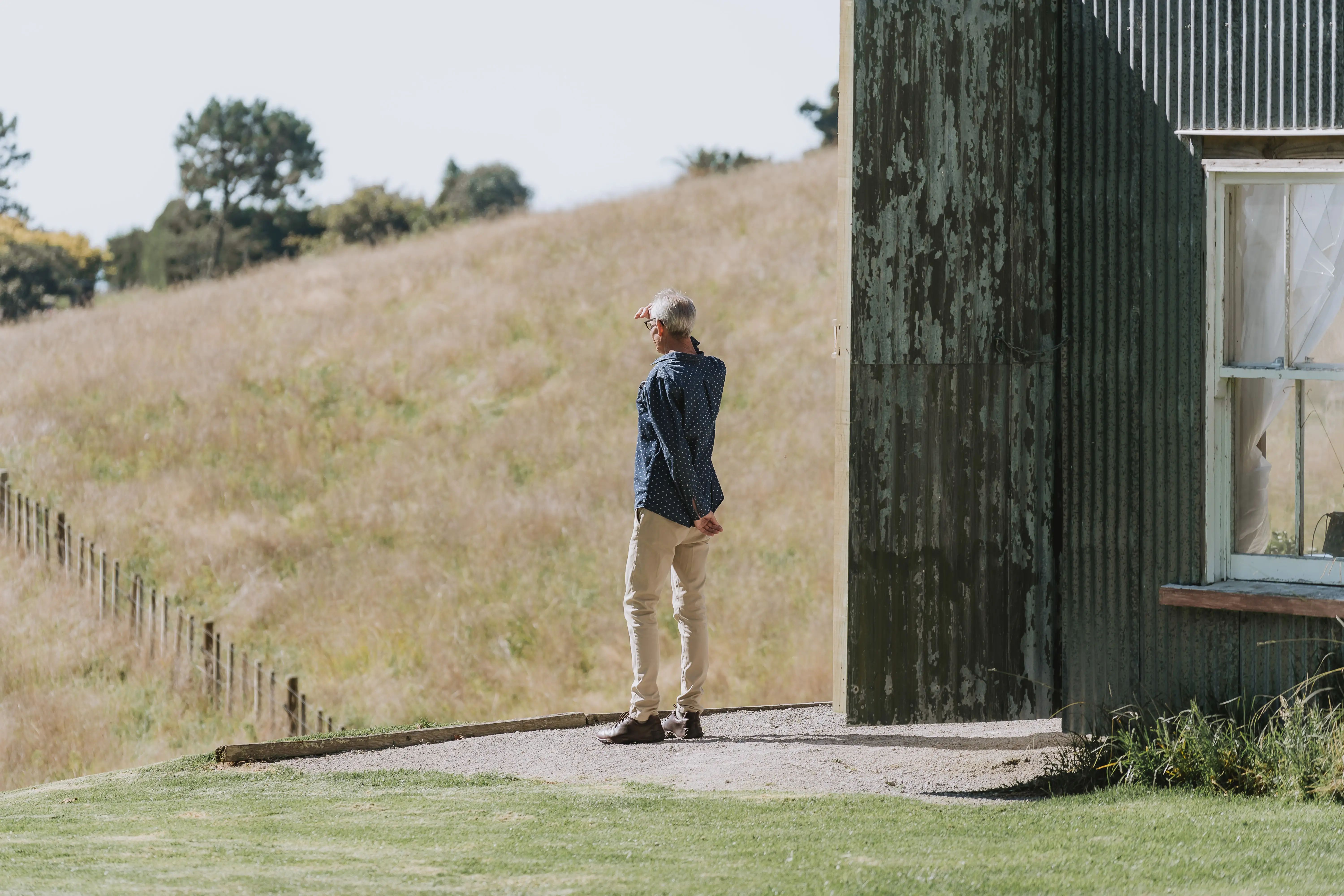 A person stands on a concrete slab near a weathered green metal building, looking into the distance with one hand shielding their eyes. Behind them is a grassy field with a slight hill and scattered trees.