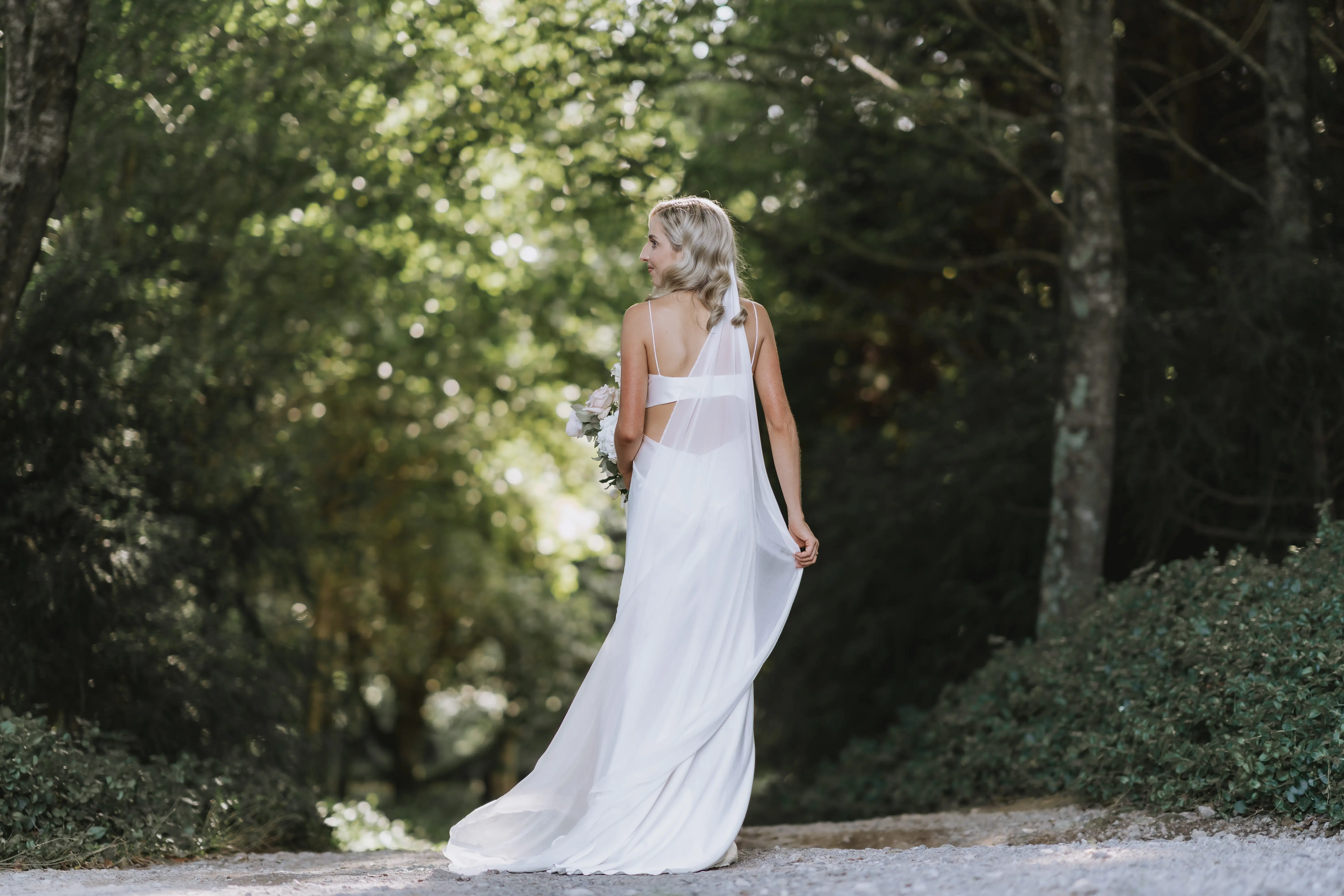 A woman in a flowing white dress stands on a path surrounded by lush green trees, holding a bouquet and looking back over her shoulder. Sunlight filters through the foliage, creating a serene, romantic atmosphere.