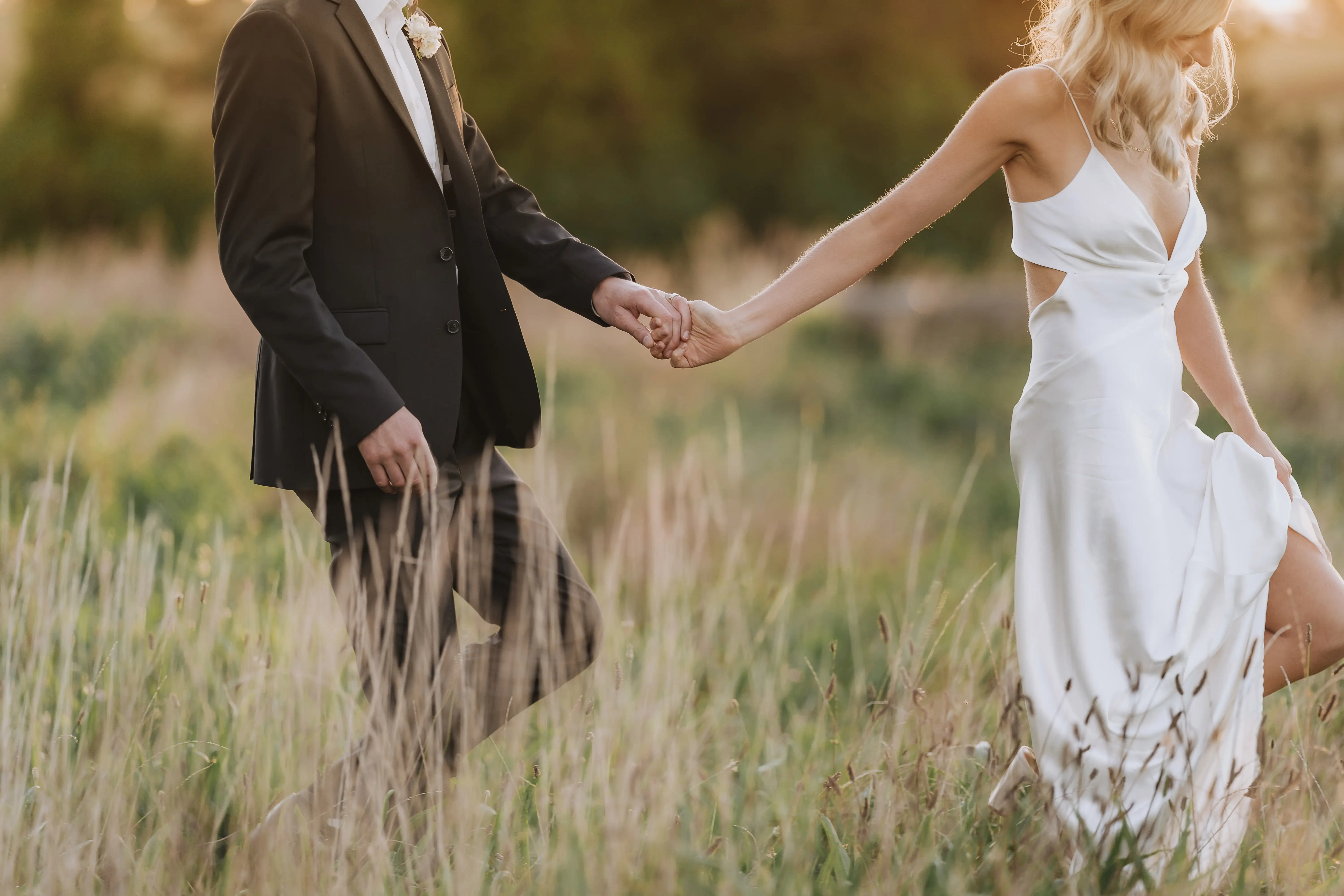 A couple dressed in formal wedding attire holds hands while walking through tall grass in a sunlit field. The woman wears a white dress and the man wears a dark suit with a boutonnière.