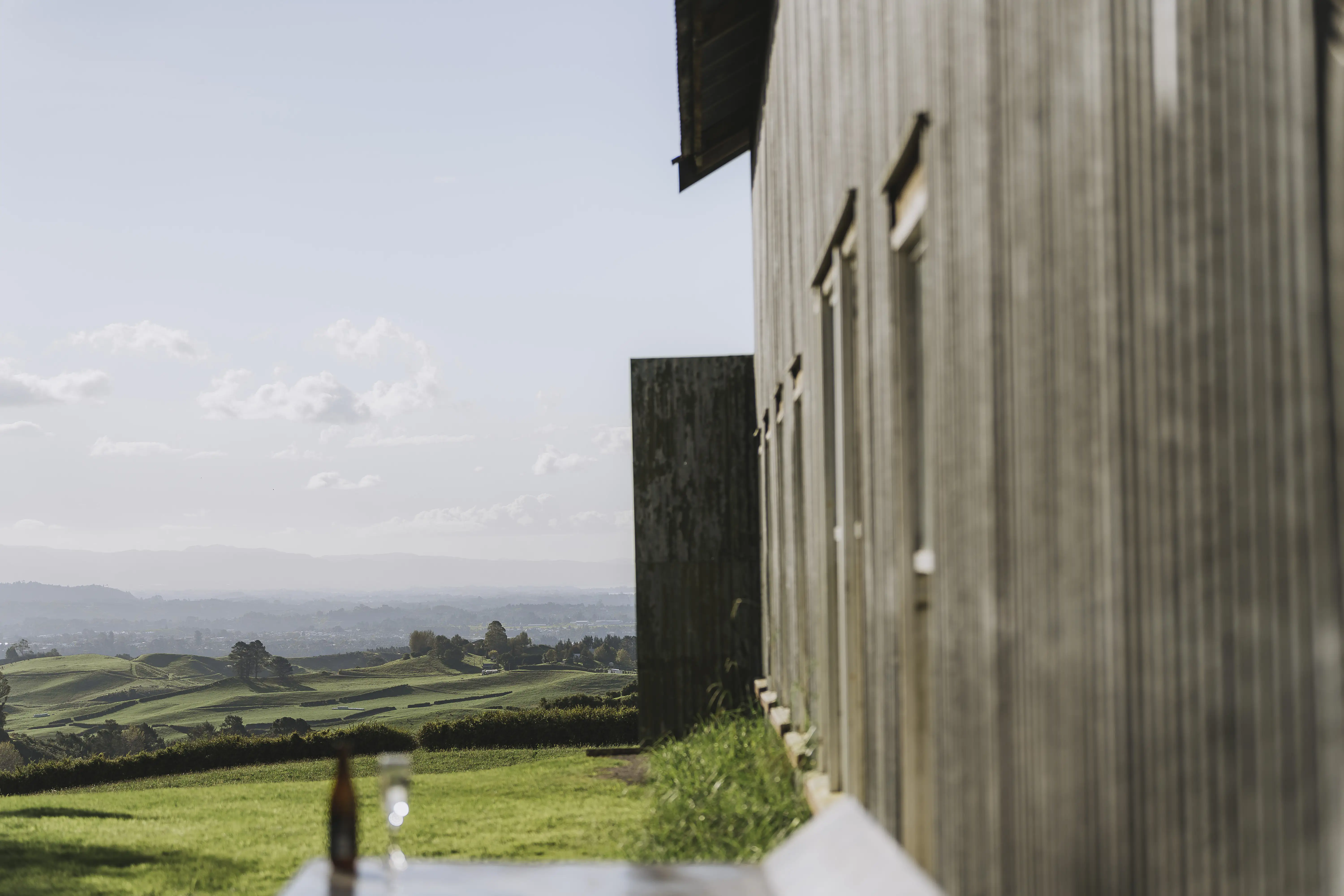 A wooden building stands on the right, overlooking green rolling hills under a blue sky with scattered clouds. A glass sits on a reflective table in the foreground. The scene is calm and sunlit.