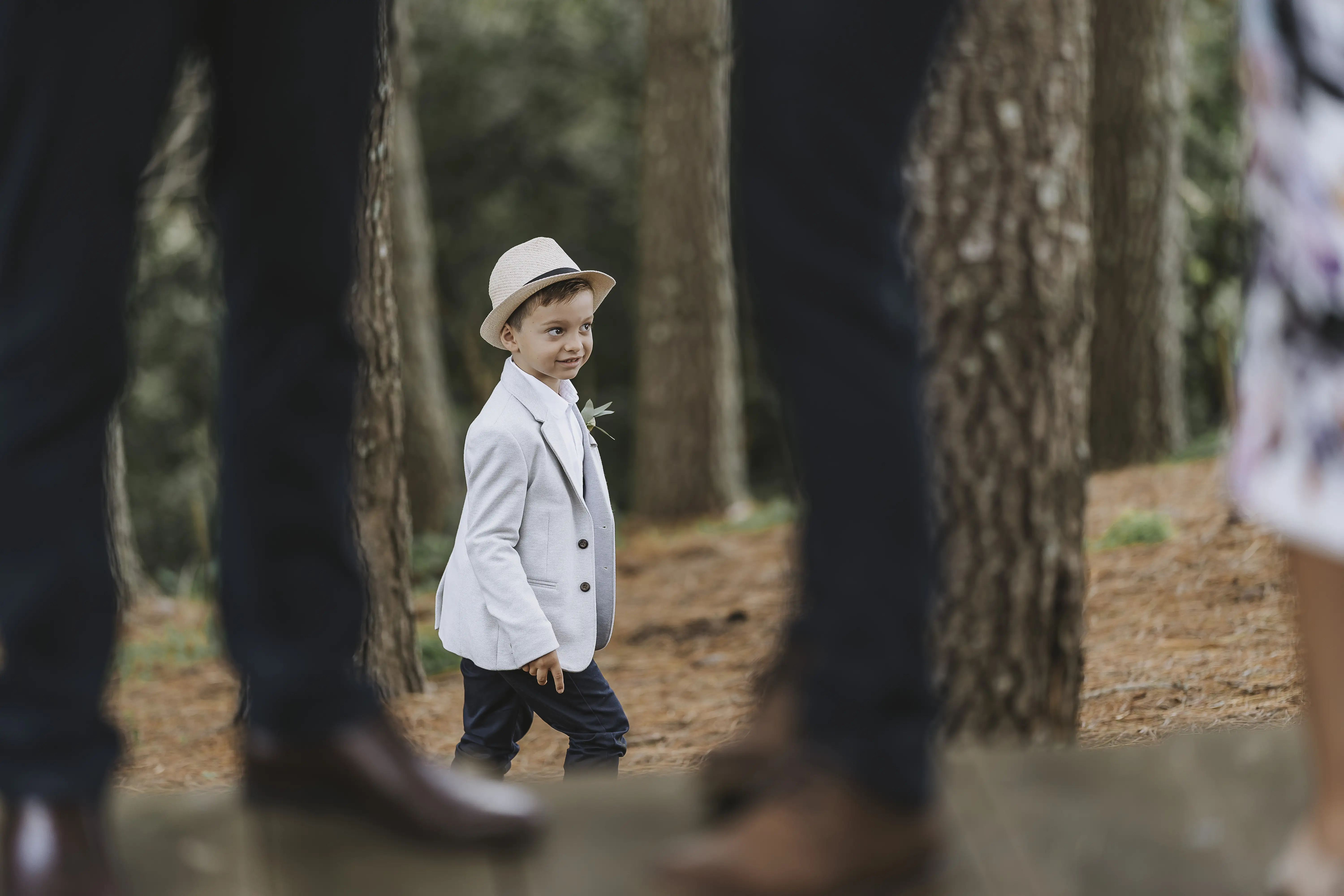 A young boy in a light blazer, dark pants, and a straw hat walks in a forest, looking up and smiling, framed by blurred adults' legs in the foreground.