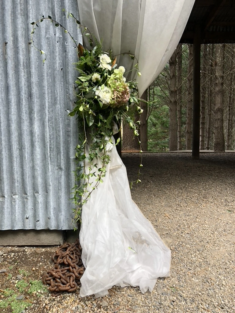A white sheer curtain is tied back with greenery and white flowers against a corrugated metal wall. The curtain pools on the gravel ground, with a rusty chain partially visible underneath. Tall trees are in the background.