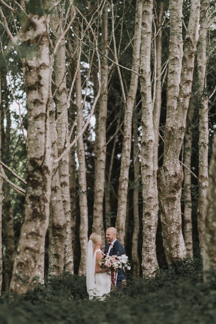 A bride and groom stand facing each other in a forest, surrounded by tall trees with light-colored bark. The bride holds a bouquet and wears a white dress and veil; the groom wears a dark suit.