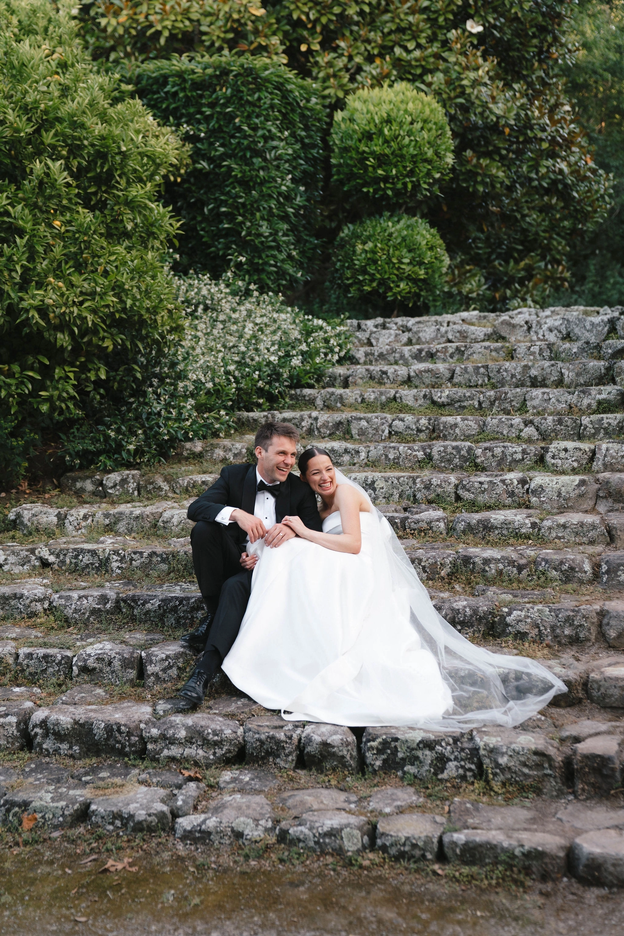 A bride and groom sit on stone steps outdoors, laughing and leaning into each other, surrounded by lush green bushes and trees. The bride wears a white dress and veil; the groom is in a black tuxedo.