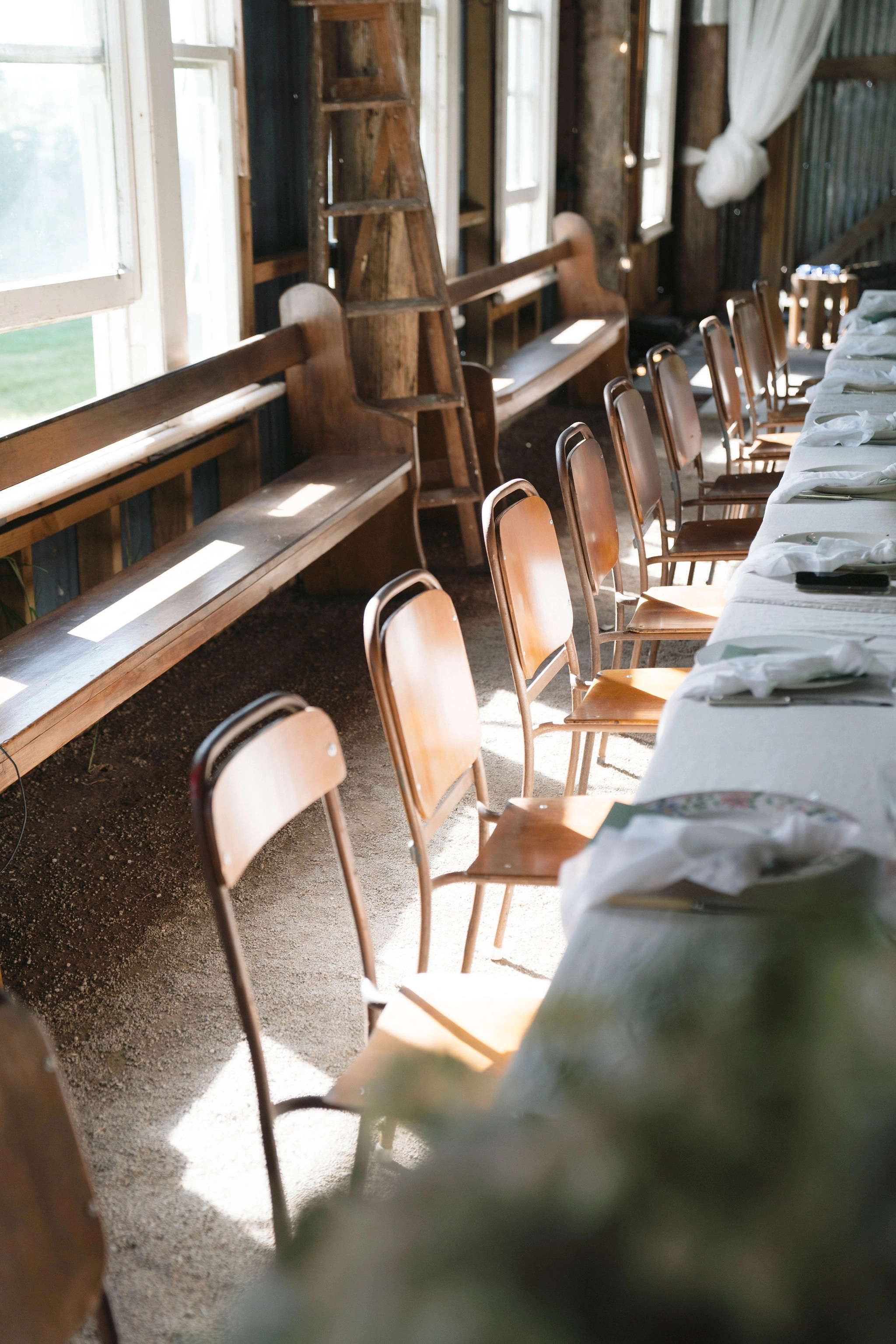Sunlight streams through large windows onto a row of empty wooden chairs and a long table set with white cloths, in a rustic room with wooden benches, a ladder, and exposed walls.