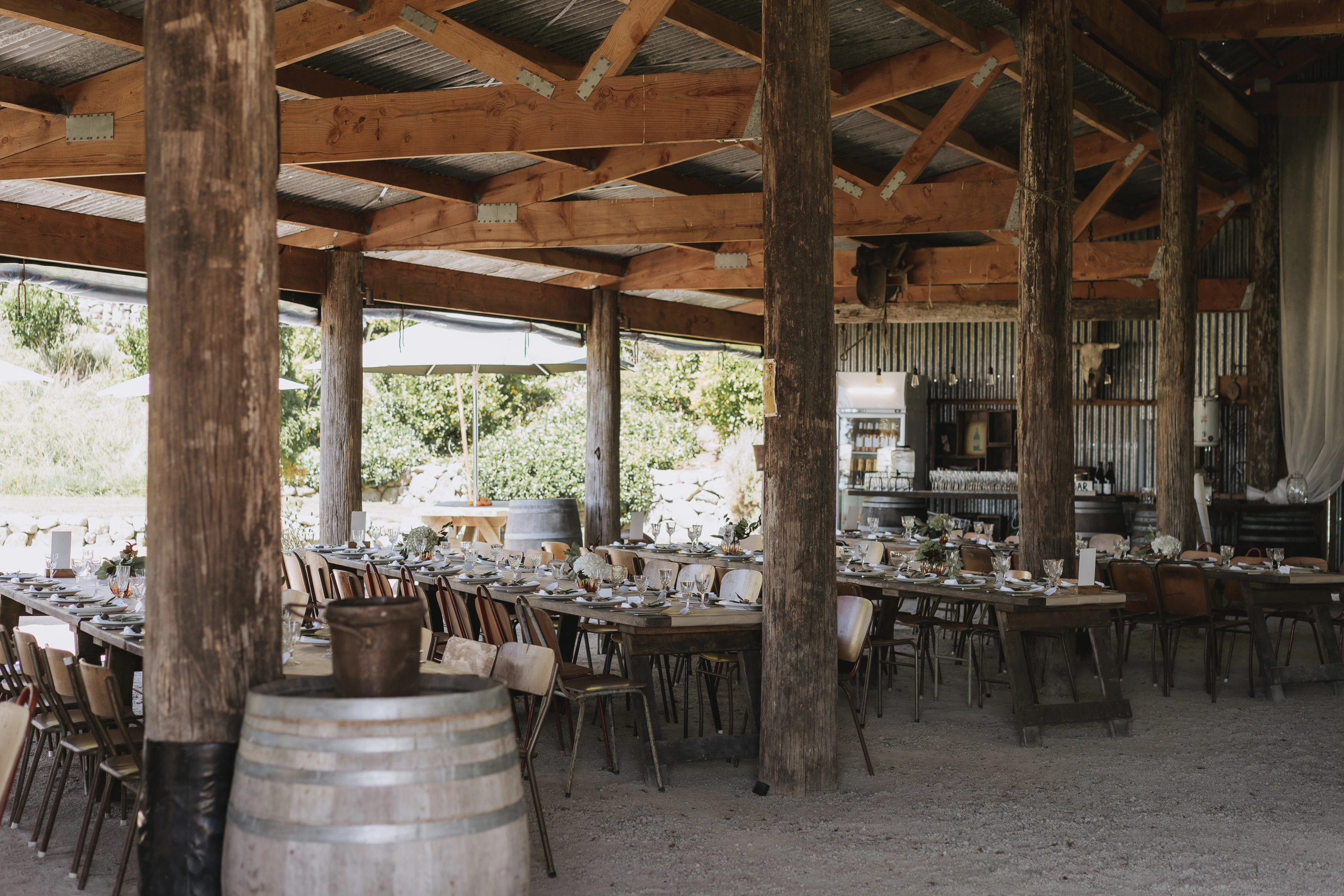 Rustic open-air barn with long wooden tables set for a meal, surrounded by mismatched chairs. Wooden beams, a barrel, and corrugated metal walls add to the country atmosphere. Natural light filters in from outside.