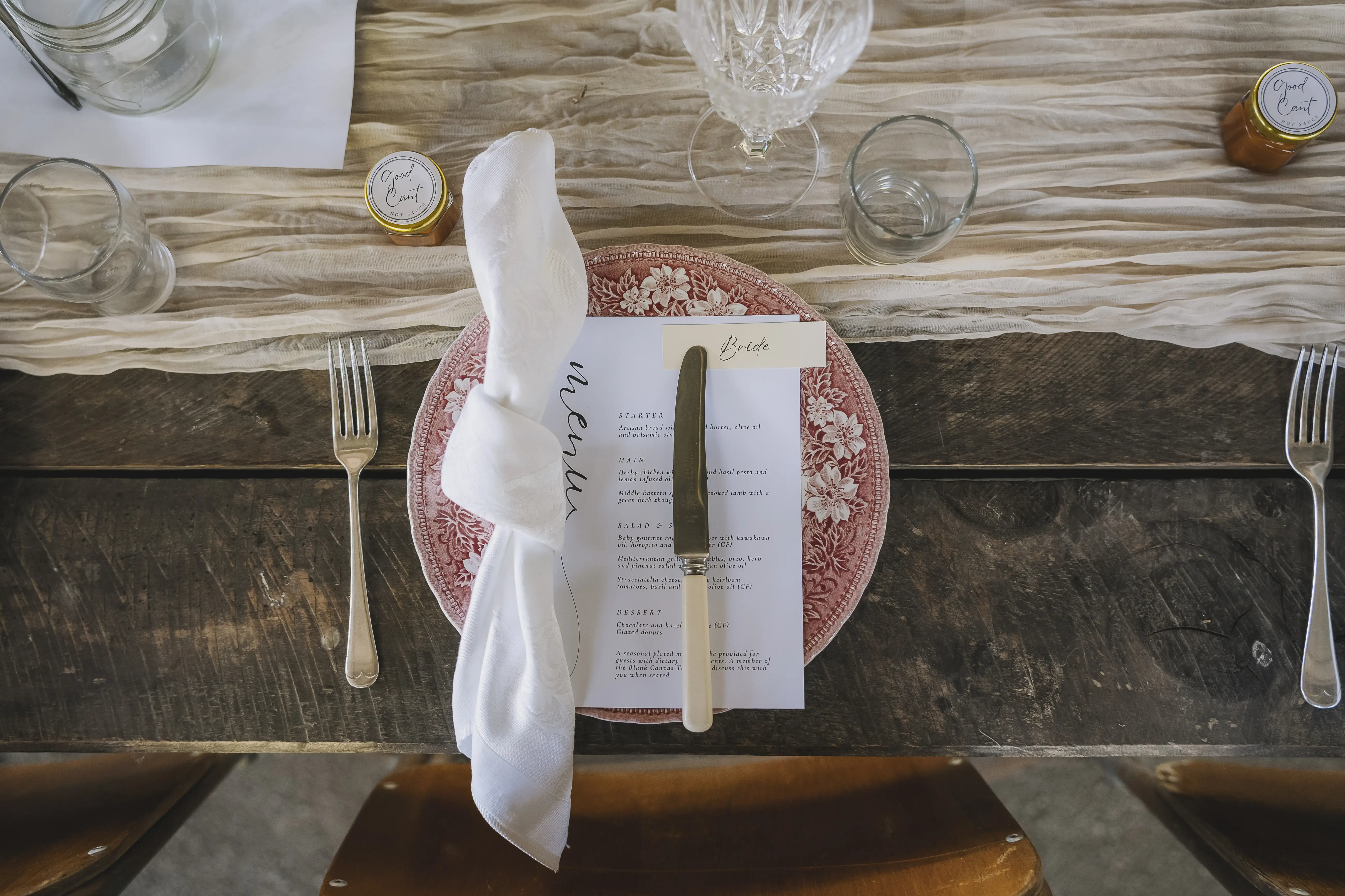 A rustic table setting with a pink patterned plate, white napkin, menu, and knife. Glassware, small jars, and simple cutlery are arranged neatly on a textured table runner over a wooden table.