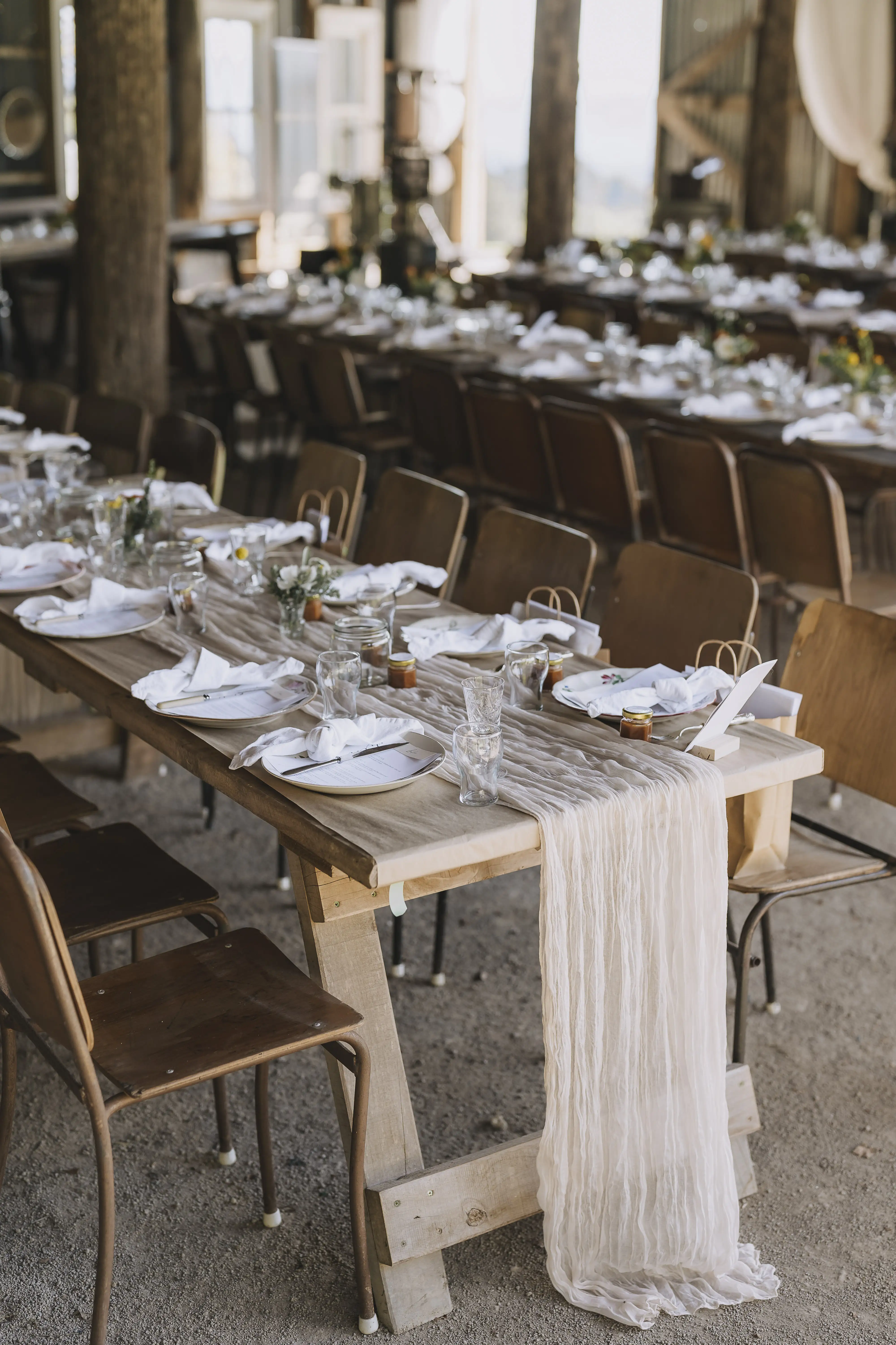 A rustic banquet table is set with white napkins, glassware, and small vases of flowers. A sheer table runner drapes down the end, and brown wooden chairs are arranged along both sides. The setting is indoors.