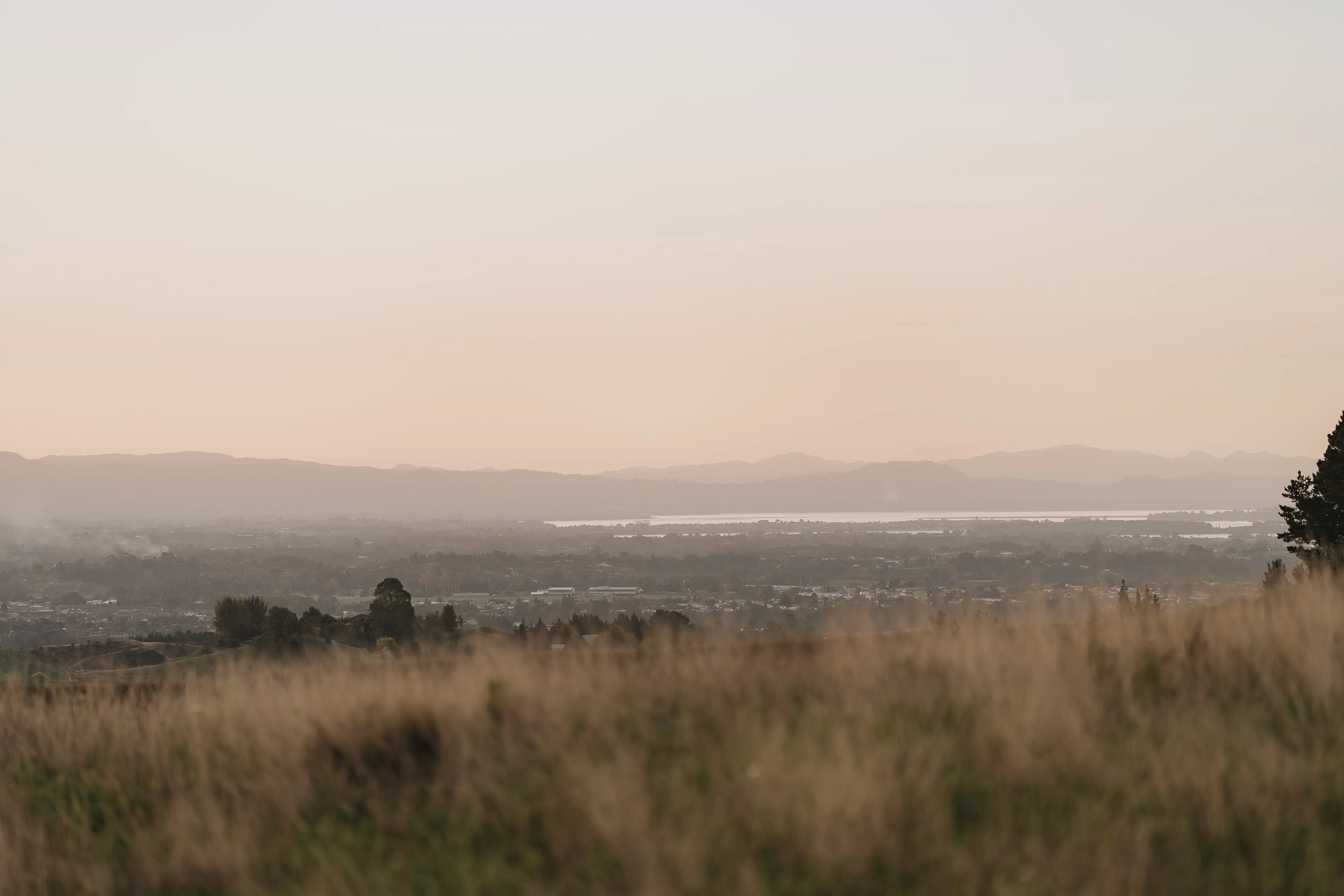 Grassy field in the foreground with a town, trees, and distant mountains under a soft, hazy sunset sky. The scene is calm and natural, with warm light and gentle colors.