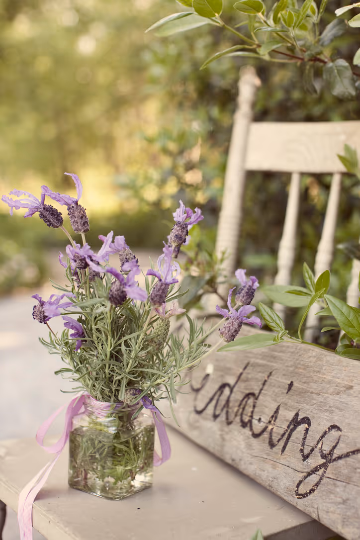 A jar with purple flowers and greenery sits on a rustic chair next to a wooden sign with "wedding" written on it, surrounded by lush green foliage and soft natural light.