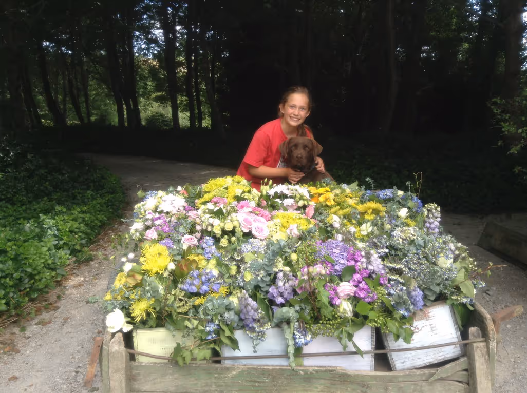 A smiling person in a red shirt hugs a brown dog behind a cart filled with colorful flowers, on a shaded path surrounded by green trees and foliage.