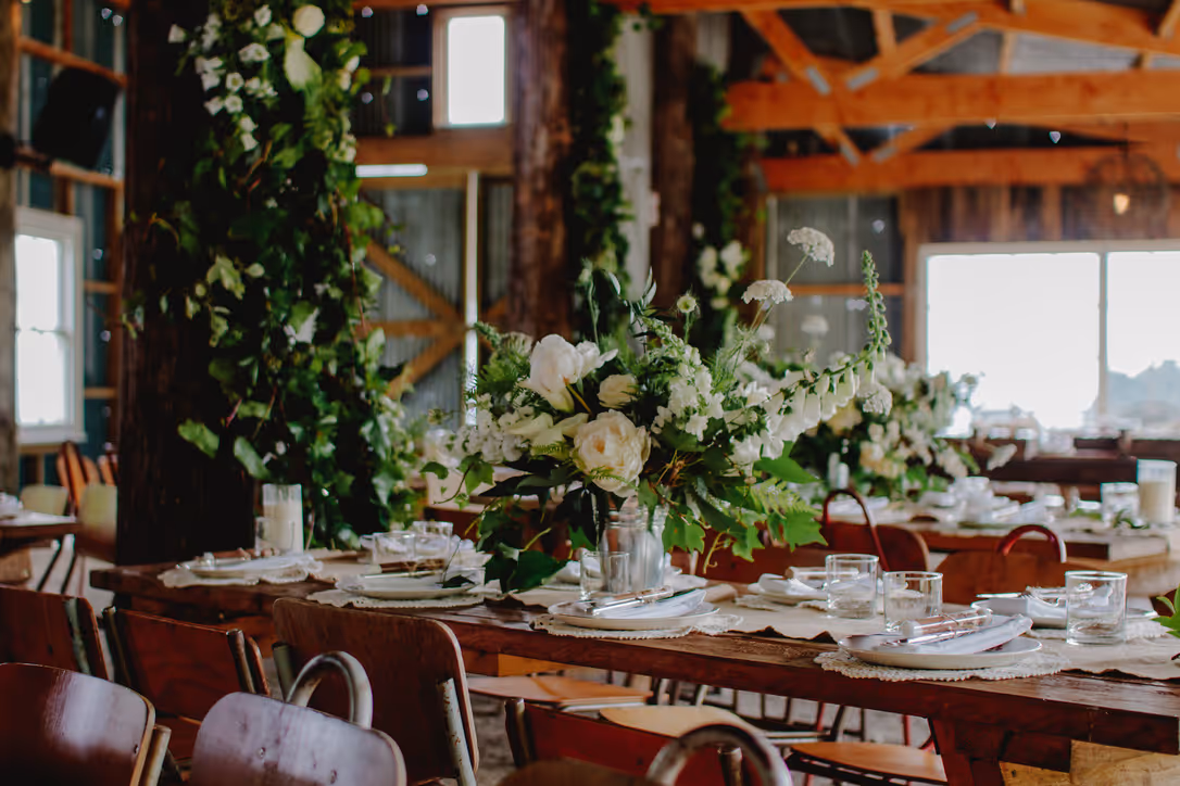 Rustic indoor event with wooden tables decorated with white flowers, greenery, candles, and glassware; wooden beams, natural light, and lush floral arrangements create an elegant, festive atmosphere.