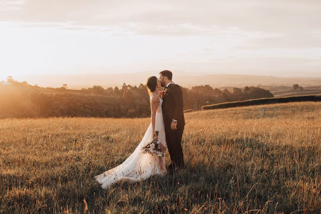 A bride and groom stand in a sunlit field at sunset, sharing a kiss. The bride holds a bouquet, and both are dressed in formal wedding attire, with rolling hills and soft golden light in the background.