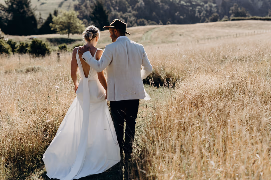 A bride in a white dress and a groom in a light jacket and hat walk arm in arm through a sunlit field with tall grass, surrounded by rolling hills and trees.