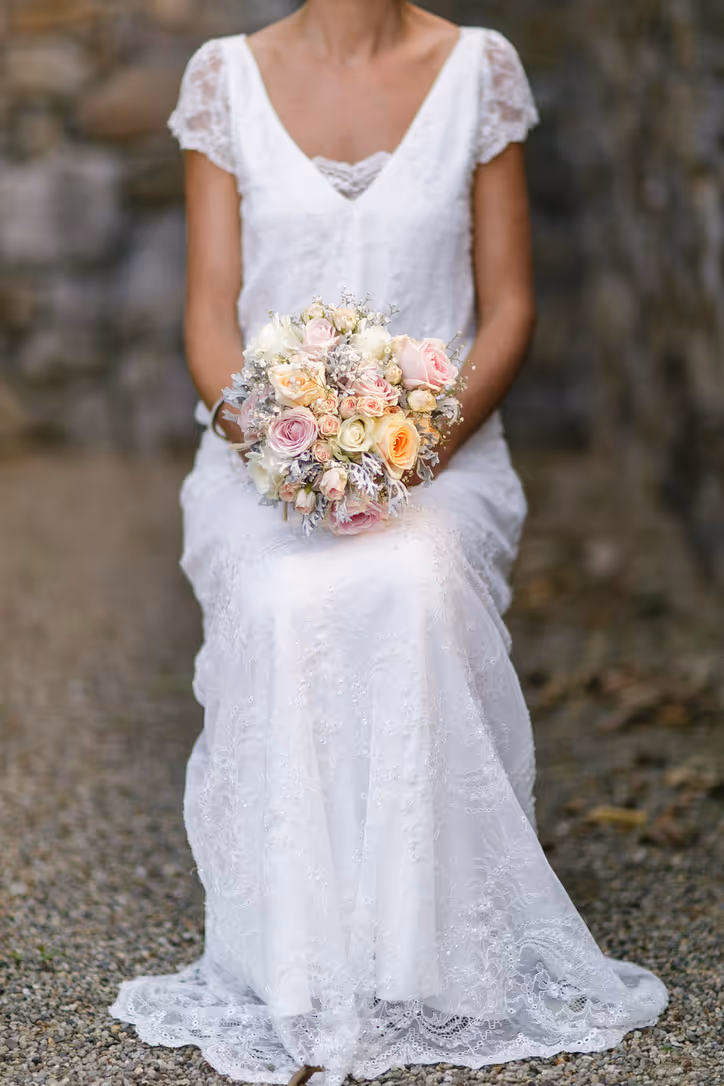 A bride in a white lace dress sits outdoors on a gravel path, holding a bouquet of pastel-colored roses and flowers. Her face is not visible in the image.