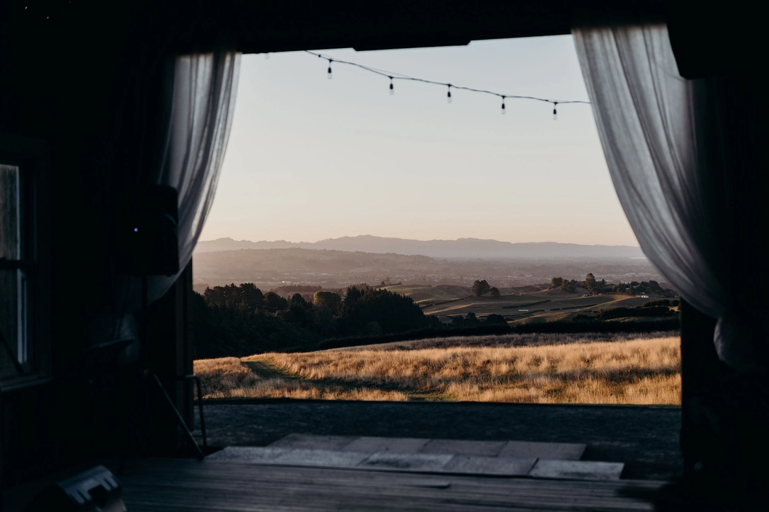 A scenic view of rolling hills and fields at sunset, seen from inside a barn with open curtains and string lights hanging above the entrance.