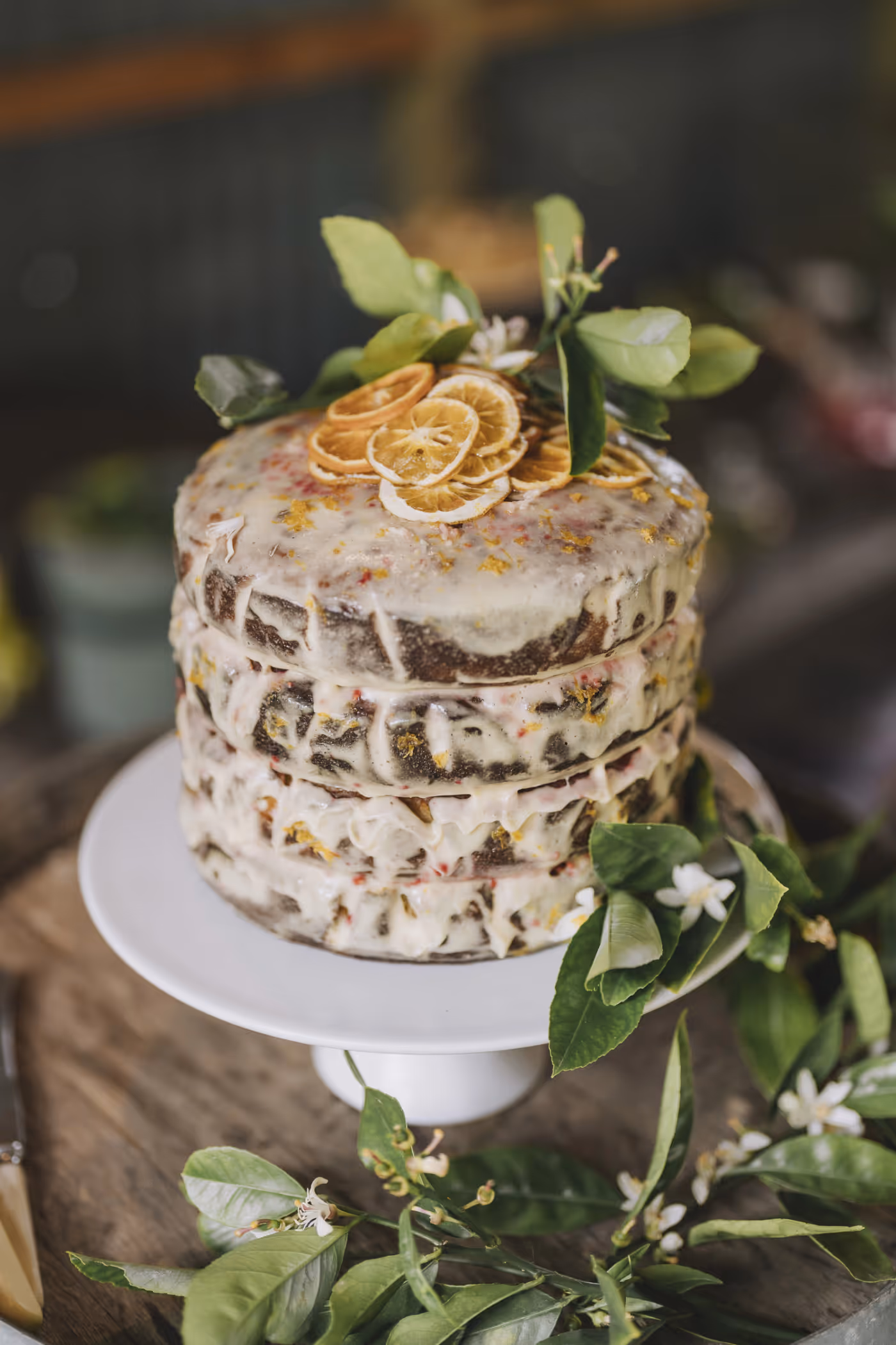 A rustic, semi-naked layer cake with white icing, decorated with dried citrus slices and fresh green leaves, sits on a white cake stand surrounded by more green foliage.