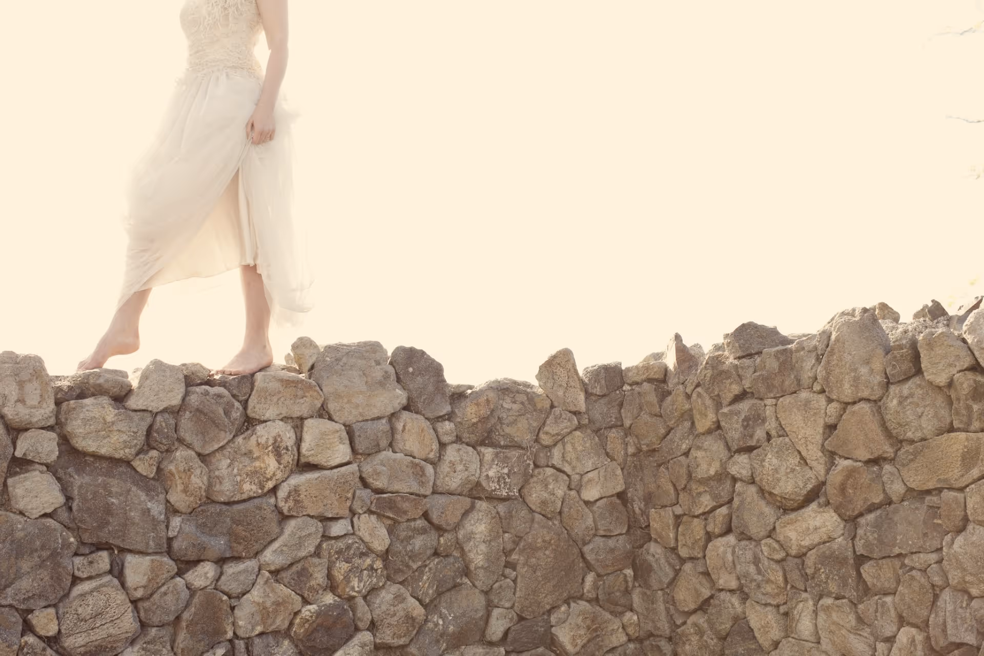 A person in a white dress walks barefoot along the top of a stone wall, with bright sunlight giving the scene a soft, warm glow. The person's head and part of their upper body are out of frame.