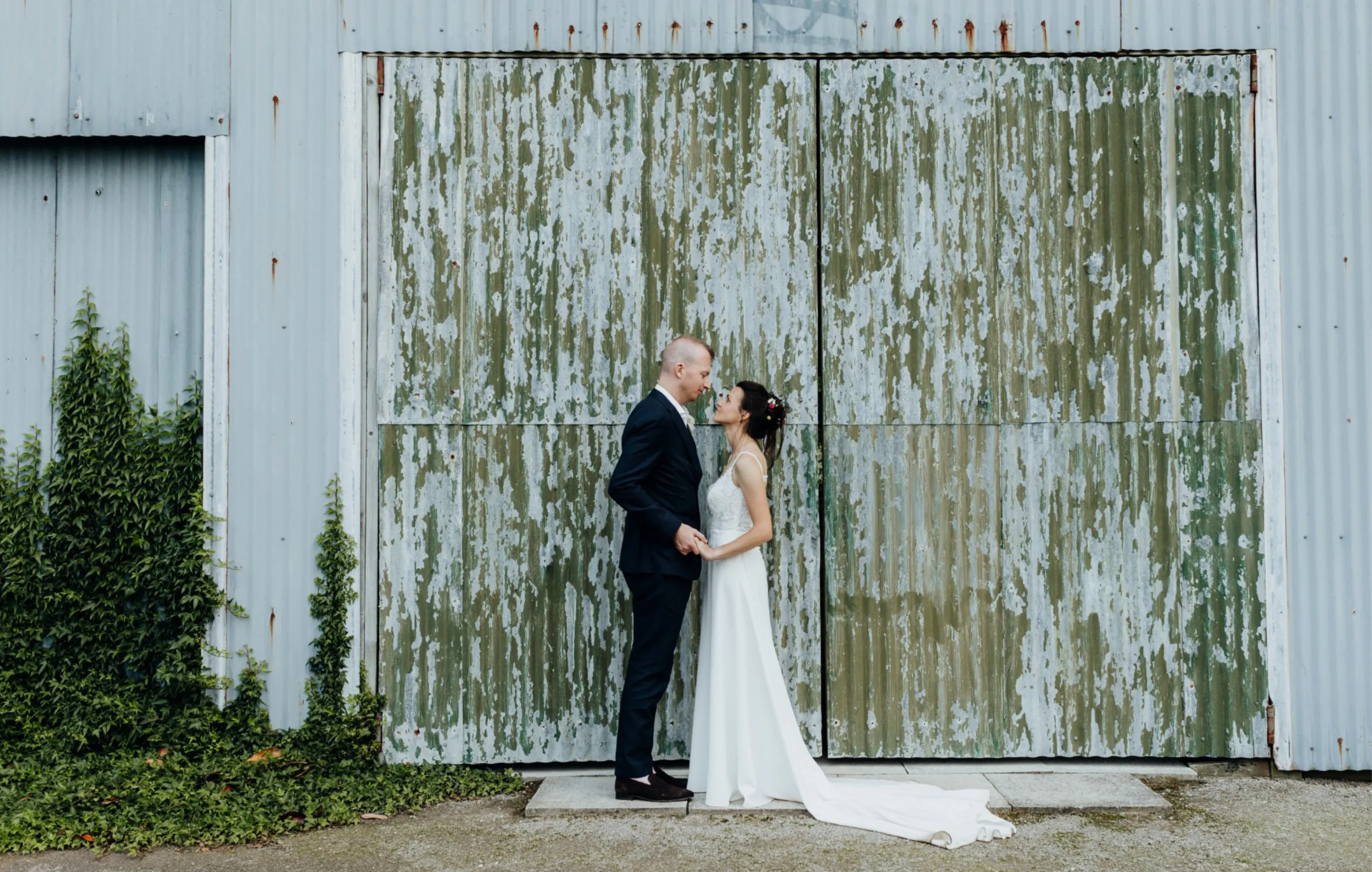 A bride and groom stand facing each other, holding hands, in front of a large, weathered metal wall with peeling paint, surrounded by a bit of ivy on the left side.