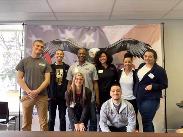 Group photo of Aldevra team members standing and smiling in front of a branded eagle backdrop in an office setting