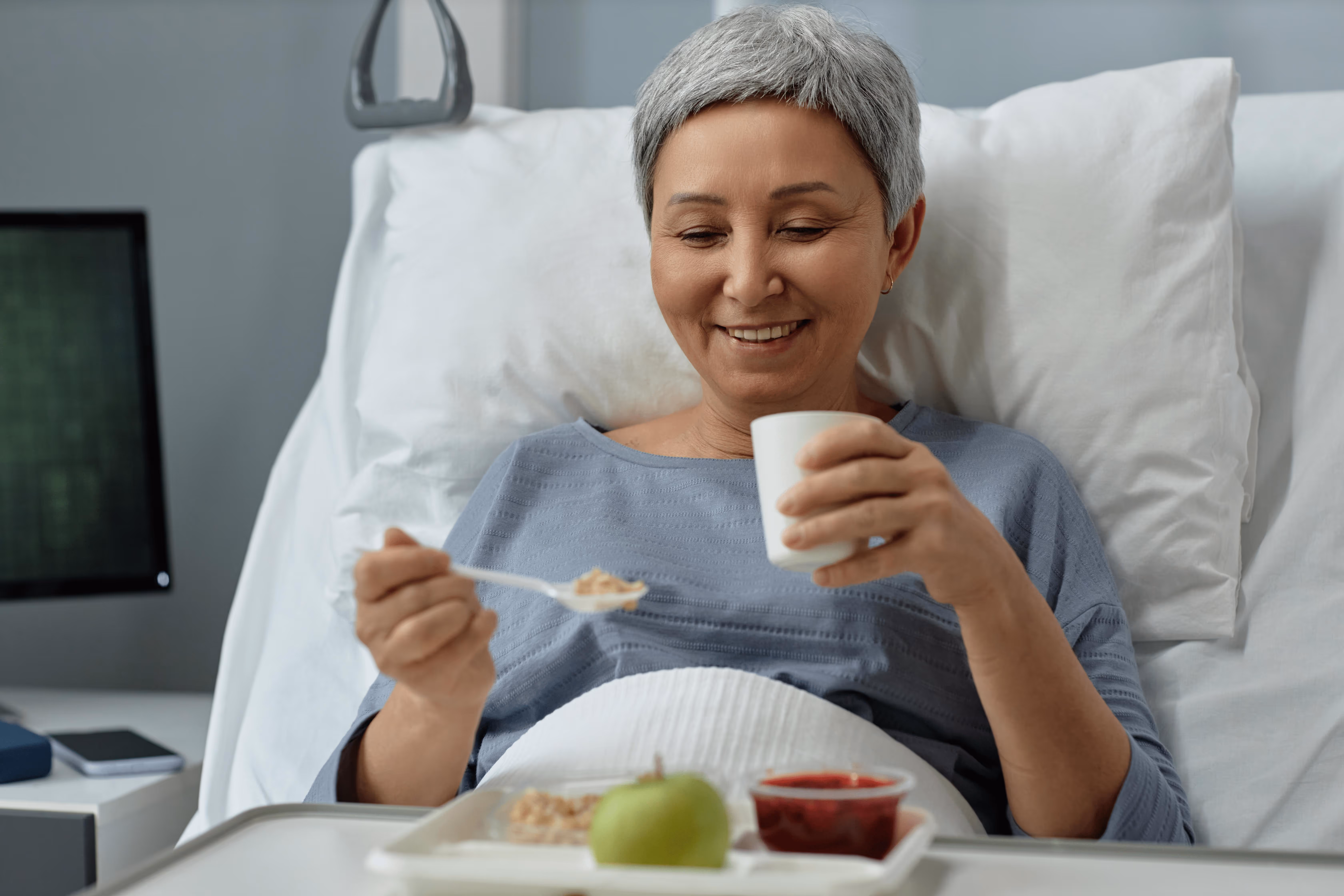 Smiling older woman with short gray hair lying in a hospital bed, holding a cup and a spoon with food on a tray in front of her.