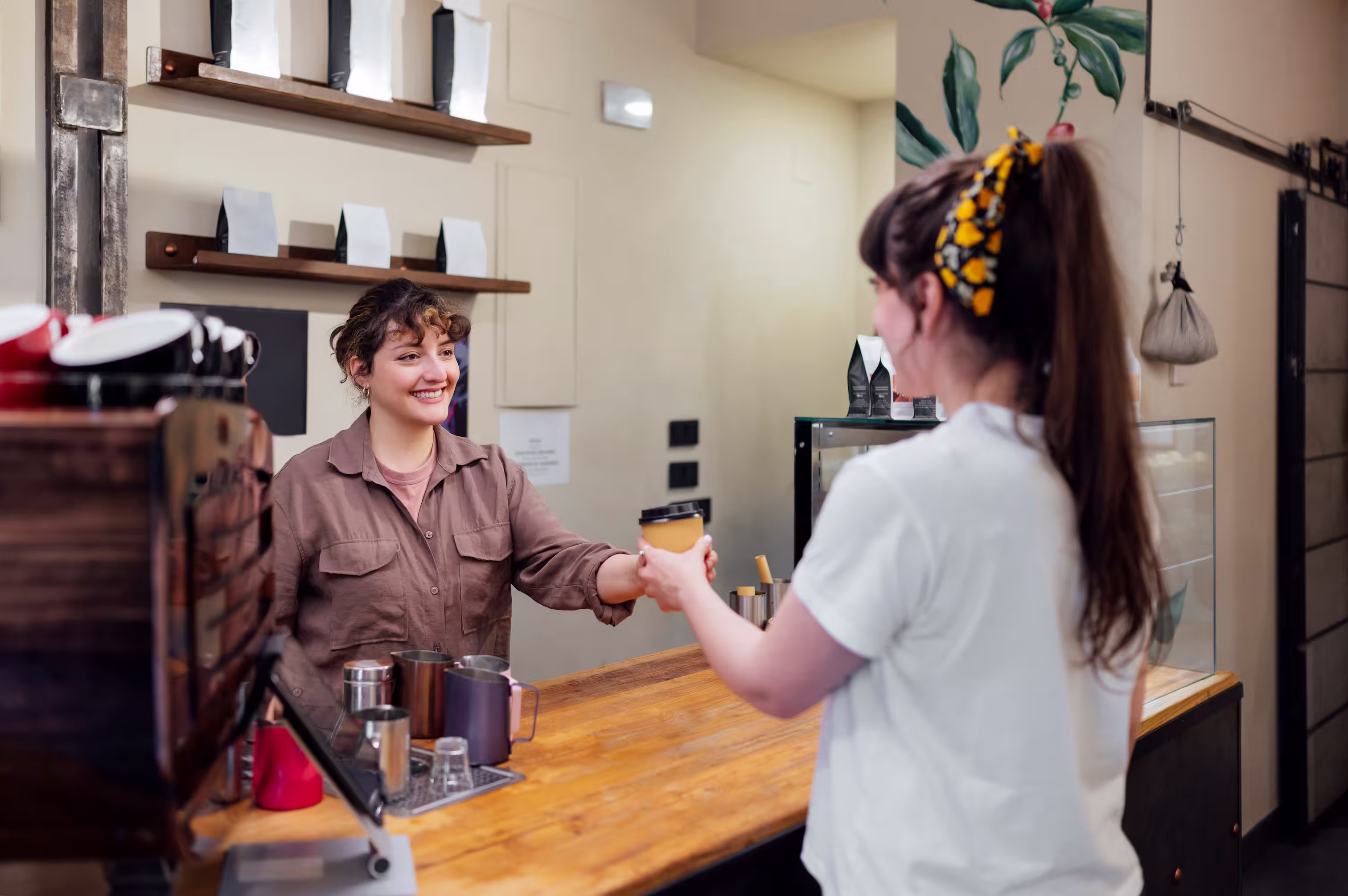 Barista smiling and handing a takeaway coffee cup to a customer at a modern coffee shop counter.