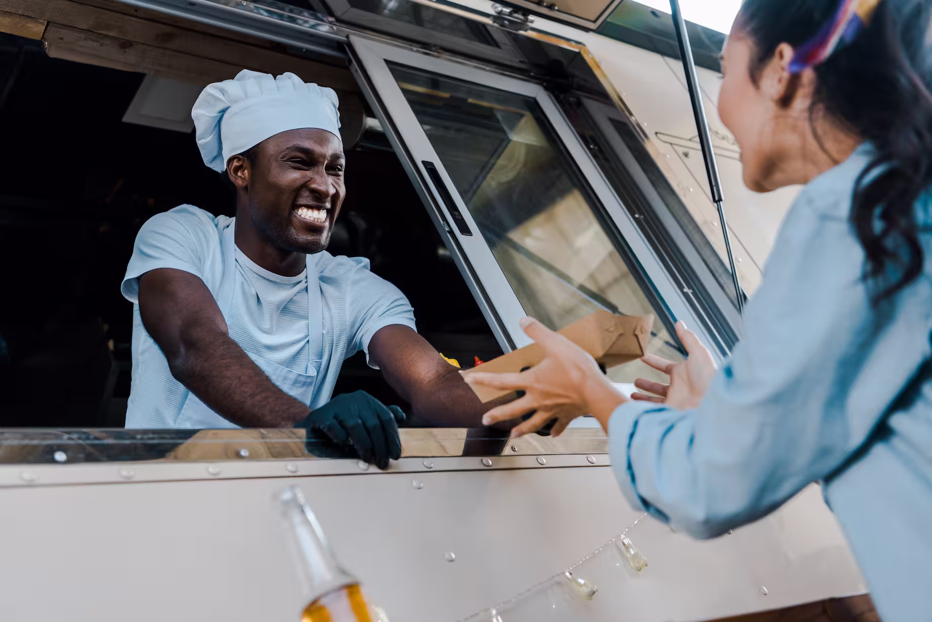 Smiling chef in a white hat handing a food package to a customer at a food truck window.