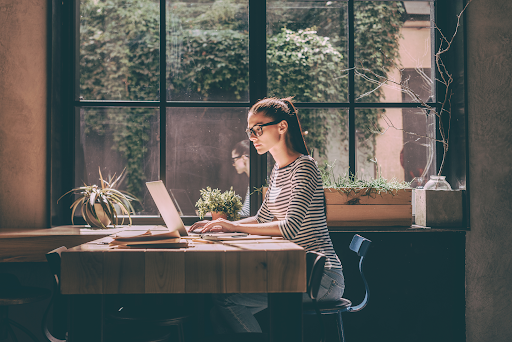 Woman working on laptop by window with plants and natural light