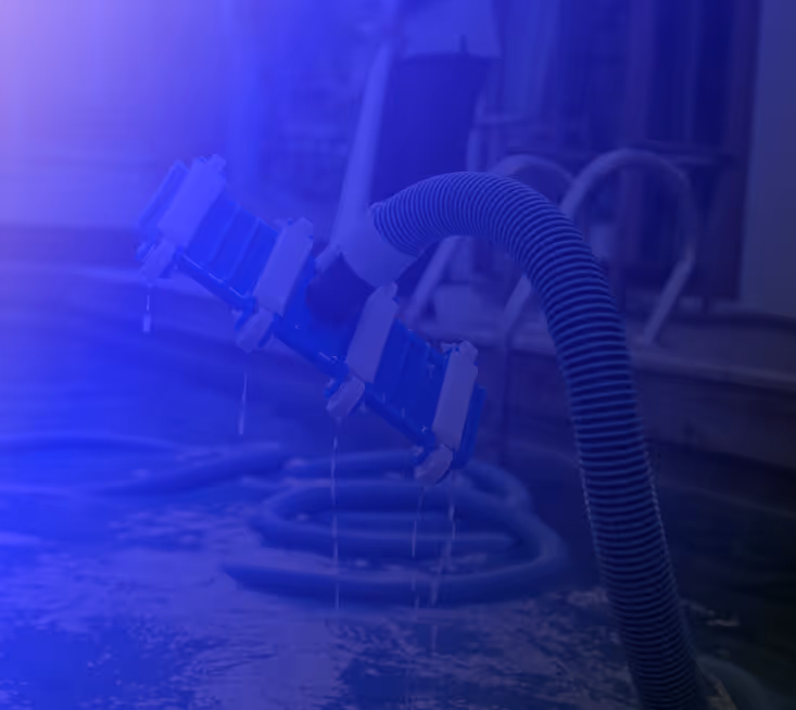Close-up of a pool vacuum system in use, with water dripping from the hose and equipment around a residential swimming pool.

