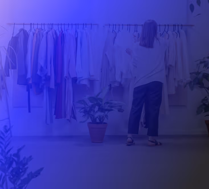 Woman browsing and organizing clothing on a retail display rack, surrounded by indoor plants in a boutique-like setting.

