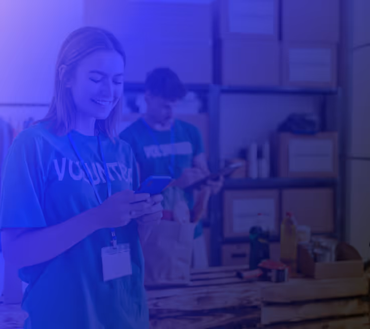 Young female volunteer smiling while using her phone in a charity storeroom, with another volunteer organizing donations in the background.

