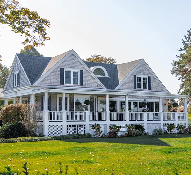 Charming shingle-style cottage with a spacious porch and lush greenery in a picturesque setting.
