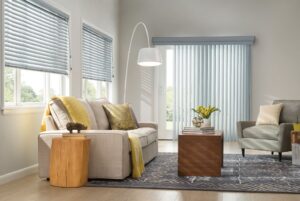 A beautiful living room with a beige couch and armchair and wood tables. There are light blue vertical blinds on the glass doors and horizontal blinds on the windows.