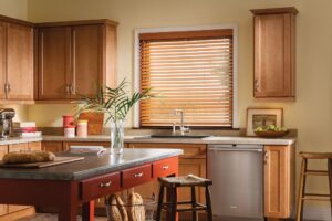 A traditional kitchen with a wood island, light wood cabinets, and a big picture window with brown faux-wood blinds.