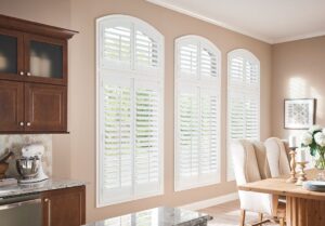 Image from the edge of a kitchen, showing part of a dining room and three big windows with white wood shutters.