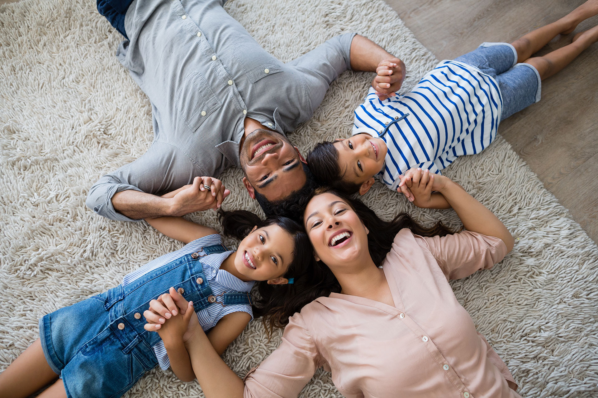 Happy family of four lying on a carpeted floor, holding hands and smiling up at the camera