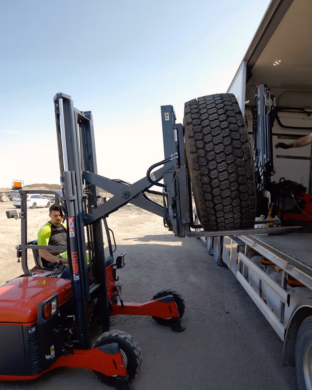 A man on a red forklift is lifting a tire out of a truck on a sunny day.