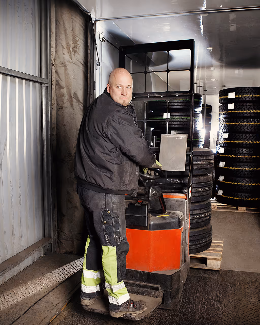 A man with black clothes drives a red fork lift carrying four tires.