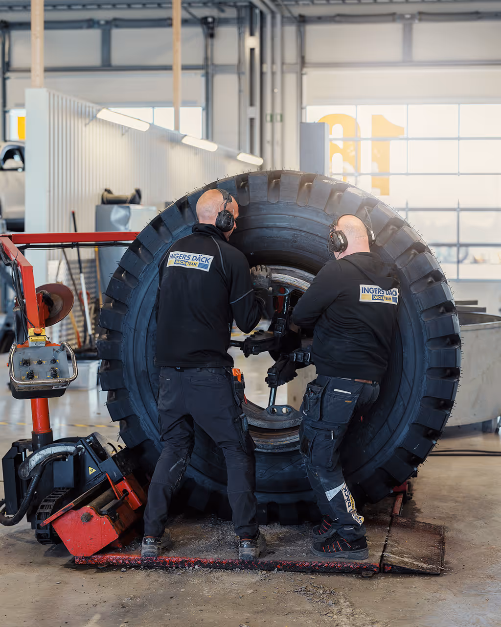 Two men are working on a big tire in a service center with large windows. They wear dark clothes with the logo Ingers Däck on their backs.