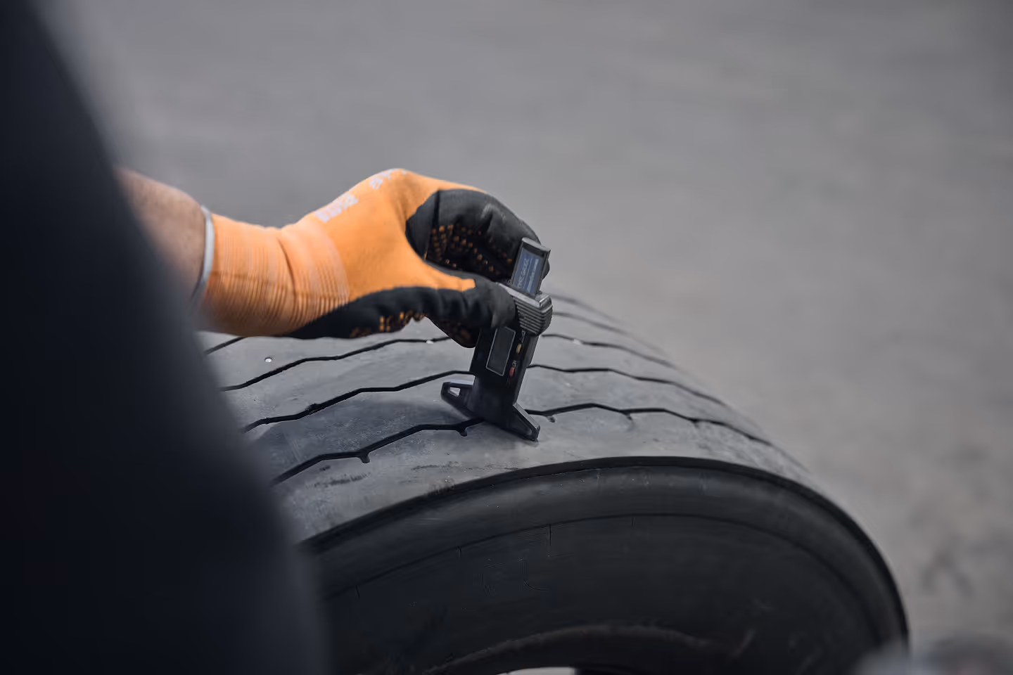 Person wearing an orange and black glove measuring tire tread depth with a digital gauge on a worn tire.