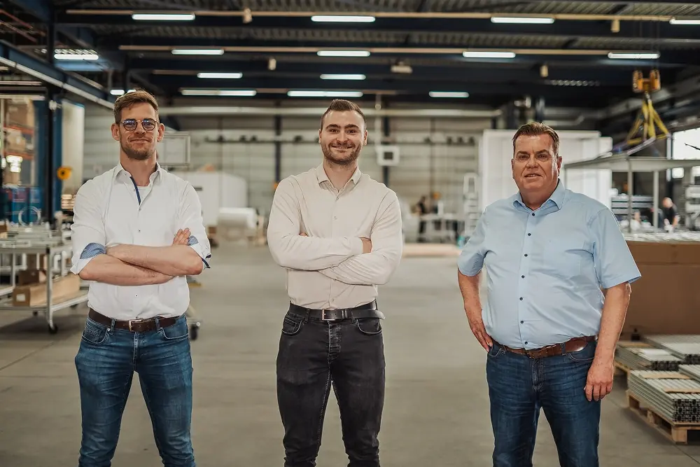 Drie mannen in een magazijn of werkplaats, staand met armen gekruist of aan de zijkant, kijkend en glimlachend naar de camera.