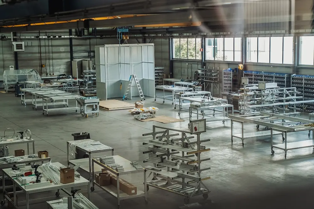 Grote industriële werkplaats met metalen werktafels, rekken en een ladder onder een hijskraan.