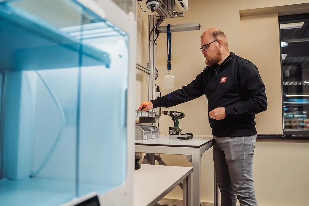 Man in werkkleding die in een werkplaats met gereedschap aan een tafel staat.