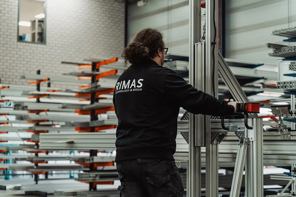 Man met zwart jack met daarop RIMAS-logo werkt aan een metalen industriële constructie in een magazijn.