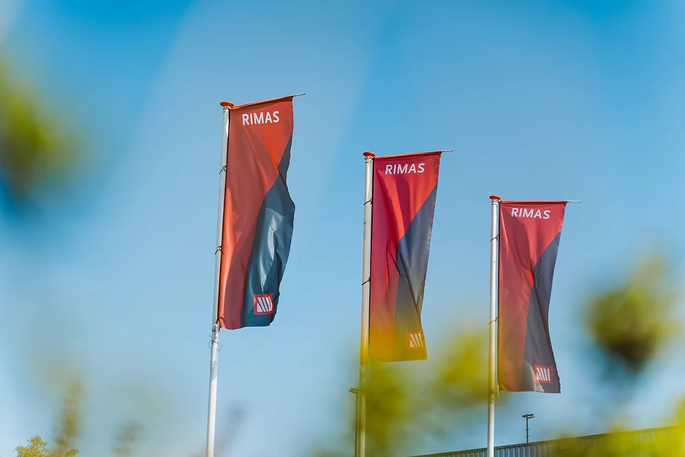 Drie rode en zwarte vlaggen op vlaggenmasten met het woord RIMAS tegen een blauwe hemel.