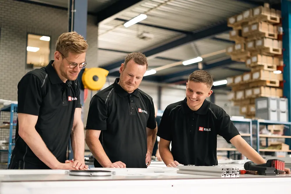 Drie mannen in zwarte Rimas-polo's bespreken iets aan een werktafel in een industriële werkplaats.