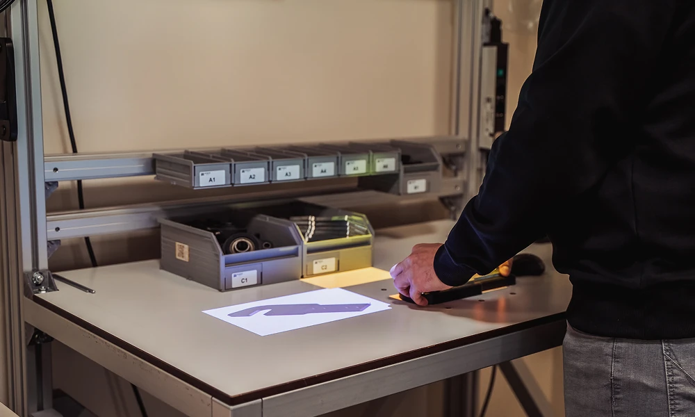 Pers die een metalen onderdeel op een industriële werkbank pakt met gelabelde materialenbakken op de achtergrond.